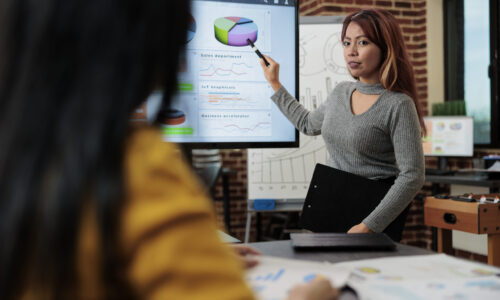 Businesswoman sitting beside monitor showing management graphs brainstorming ideas for partnership project. Marketing team discussing company strategy working in startup office. Modern workplace